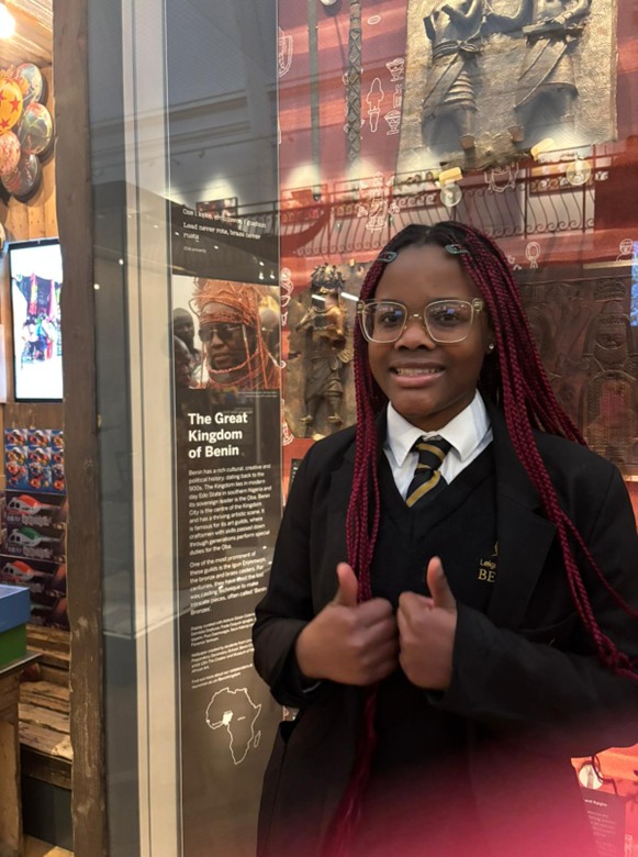 A student in a school uniform and glasses gives a double thumbs-up while standing in front of a museum exhibit titled "The Great Kingdom of Benin."