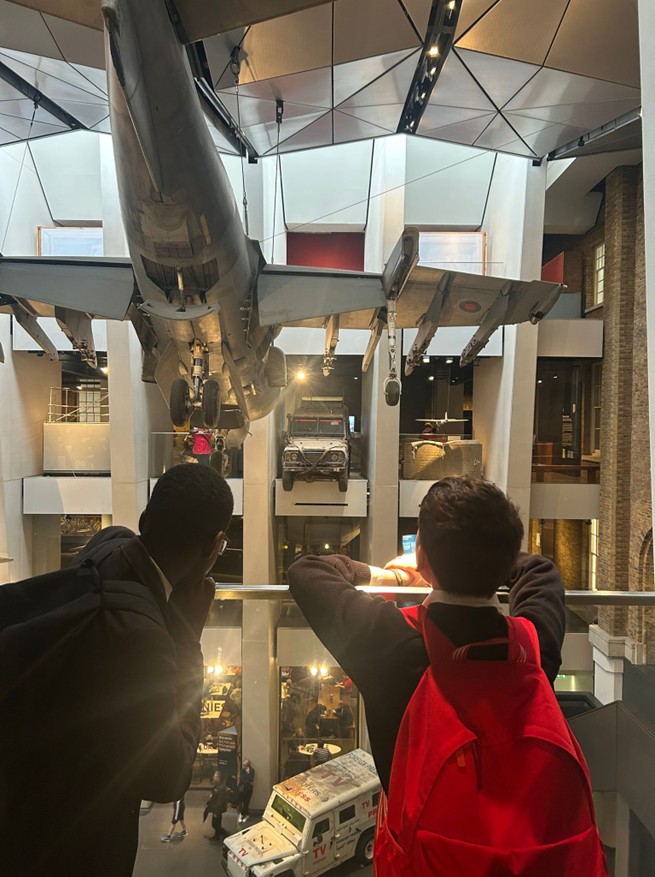 Two students lean over a railing in a museum atrium, looking at a military jet and a Land Rover suspended from the ceiling.