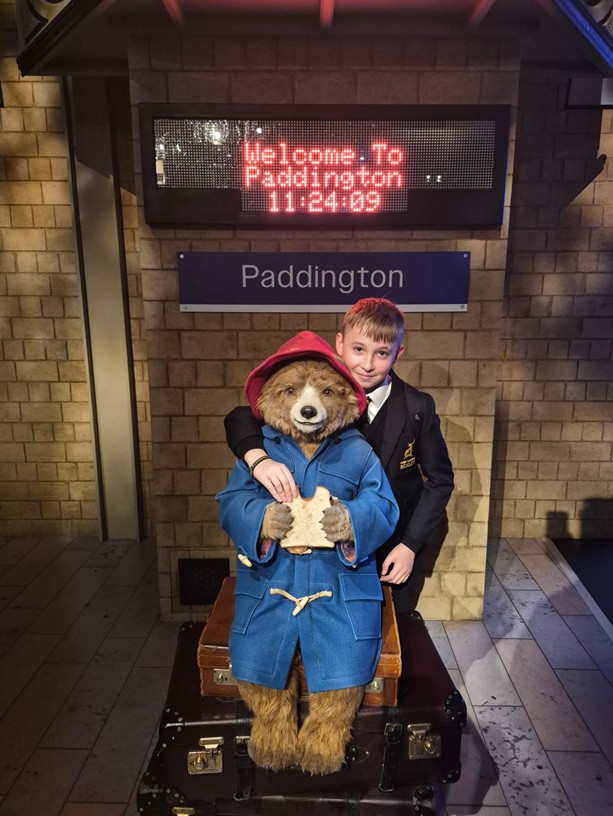 A student in a school uniform stands next to a statue of Paddington Bear at a simulated "Paddington" train station platform.