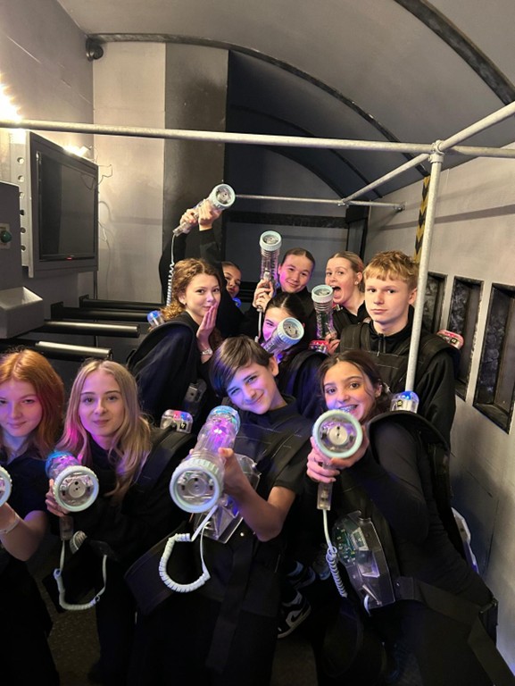 A group of students in a dimly lit, industrial-style room hold up glowing prop blasters while wearing vests, appearing to be at a laser tag or immersive experience.