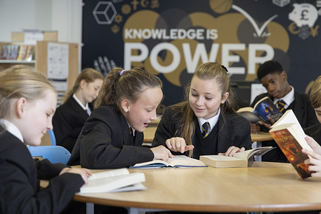 Six pupils in a classroom sitting at desks reading books. In the background on the wall it says Knowledge is power
