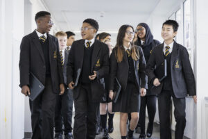 Around 12 pupils walking towards the camera in a school corridor. They are holding laptops while chatting and smiling