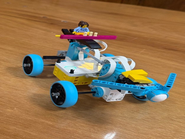 A close-up shot of a small robotic toy car assembled from blue, white, and yellow building blocks, featuring a tiny toy figure seated on top.
