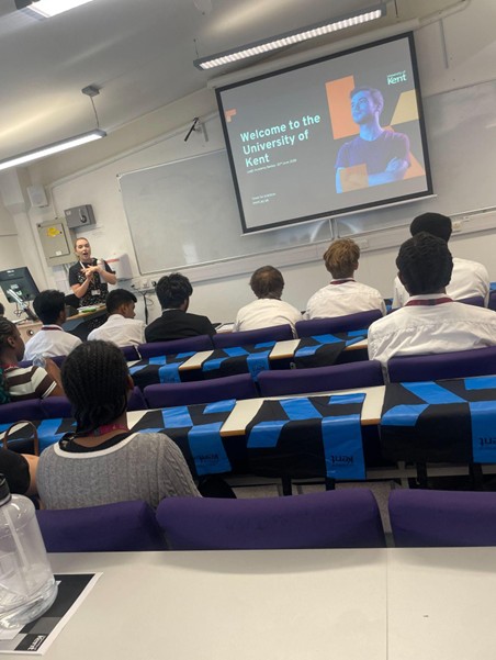 A classroom or lecture hall where a presenter stands at the front next to a projector screen displaying "Welcome to the University of Kent." Students are seated in rows of purple chairs, viewed from behind, watching the presentation.