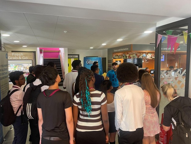 A group of students gathered inside a campus lobby or visitor reception area, listening to a guide near a "Tickets and Information" desk.