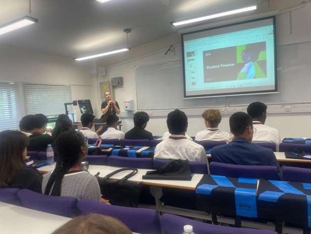 Students seated in a university lecture hall watching a presentation on "Student Finance" displayed on a projector screen at the front of the room.