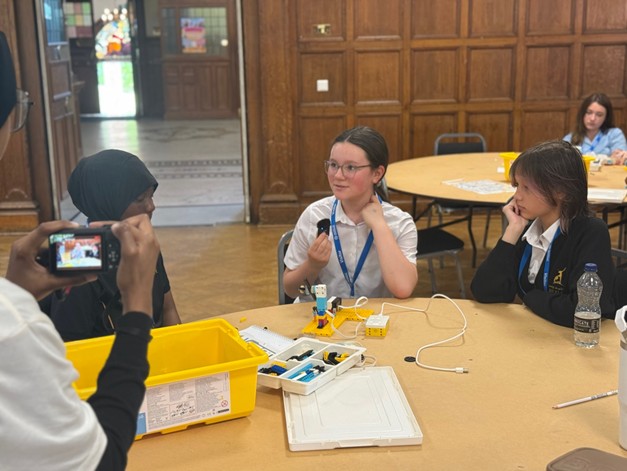 Three students sitting around a circular table working on an electronics project, while an individual in the foreground captures the moment with a digital camera.