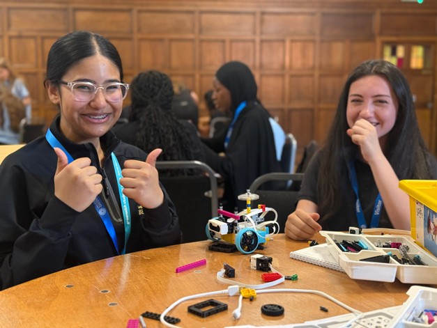 Two smiling students at a wooden table posing with a small, custom-built robotic car; one student is giving a double thumbs-up.