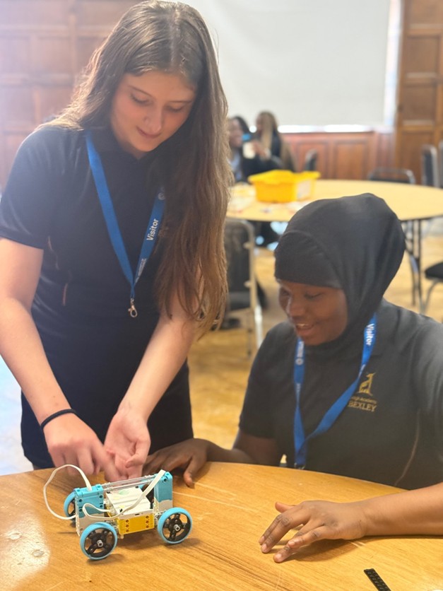 Two students collaborating closely at a table to adjust or configure a small motorized wheeled robot.