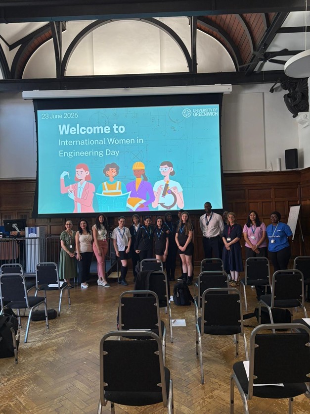 A group of individuals posing together on a stage in front of a large presentation screen that reads "Welcome to International Women in Engineering Day" at the University of Greenwich.