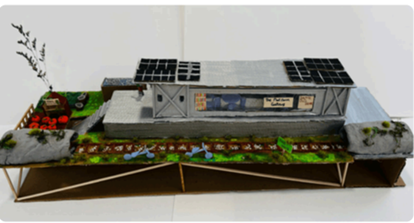 A detailed, handmade architectural model of a sustainable building featuring solar panels on the roof, a miniature garden patch, and a track running along the front.